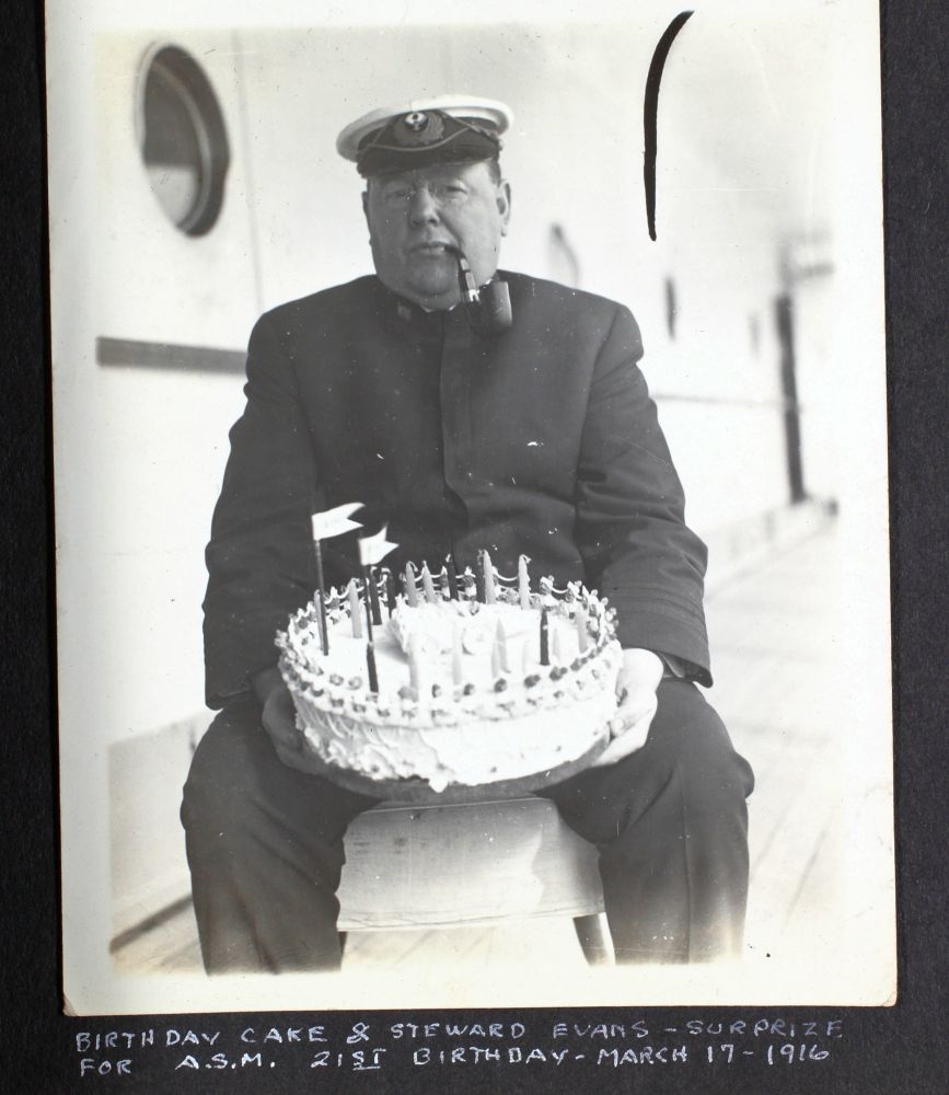 a heavyset man in a sailor outfit sitting on the deck of a ship with a pipe in his mouth. There is a largebirthday cake in his lap