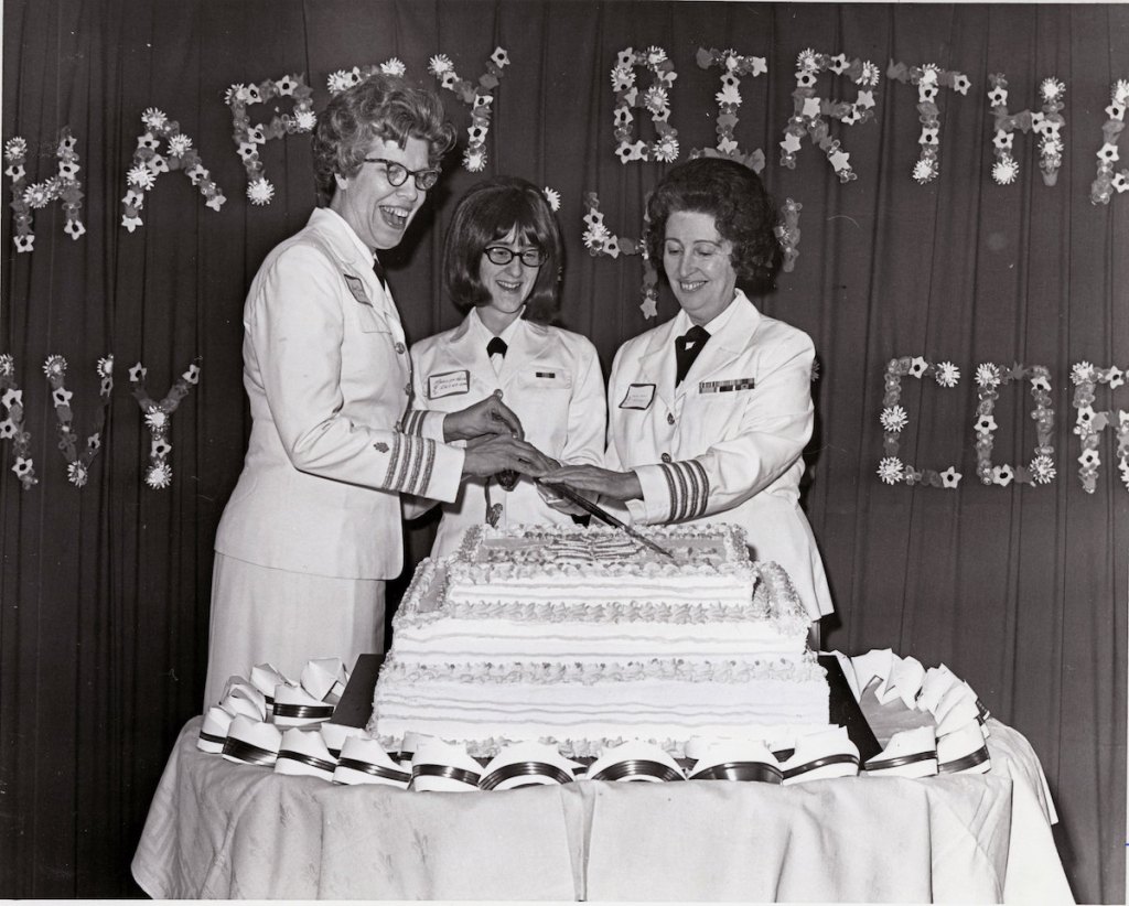 Three women cut a very large birthday cake. The words Happy Birthday are in a floral arrangement on the wall behind them