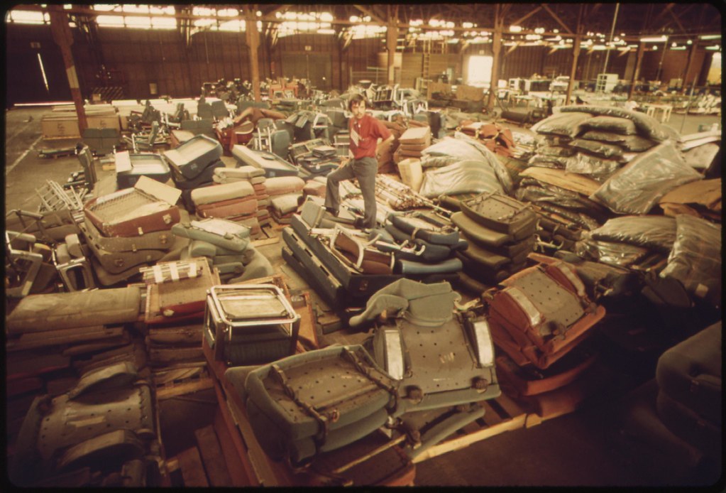 A man standing on top of some dissassembled furniture inside a warehouse full of such furtniture