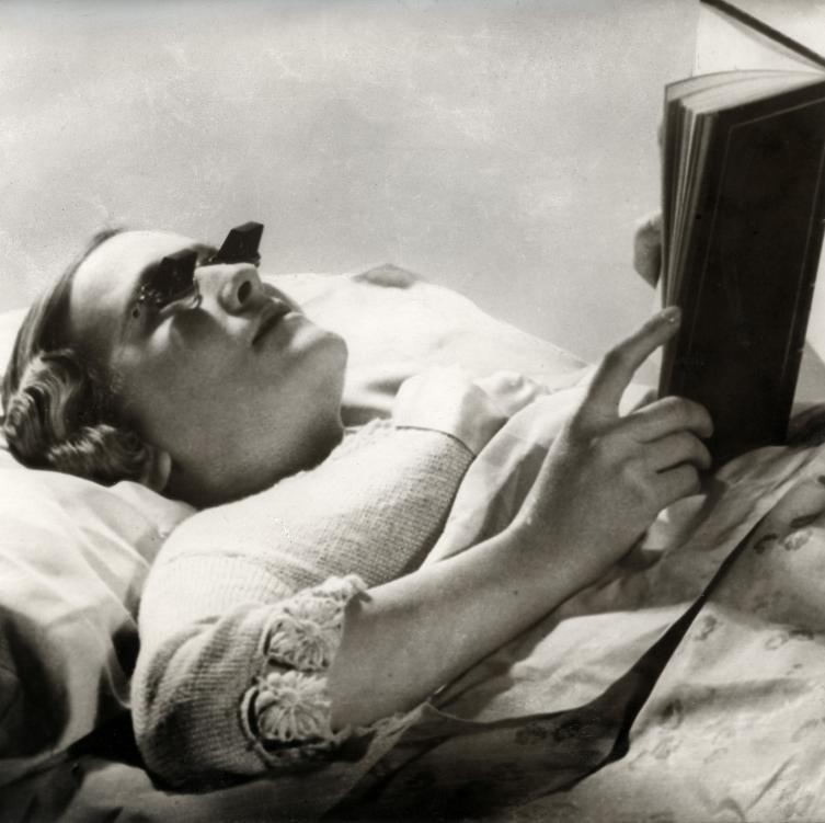 White woman lies on a bed, wearing glasses designed for reading in bed. She is resting a book on her torso.