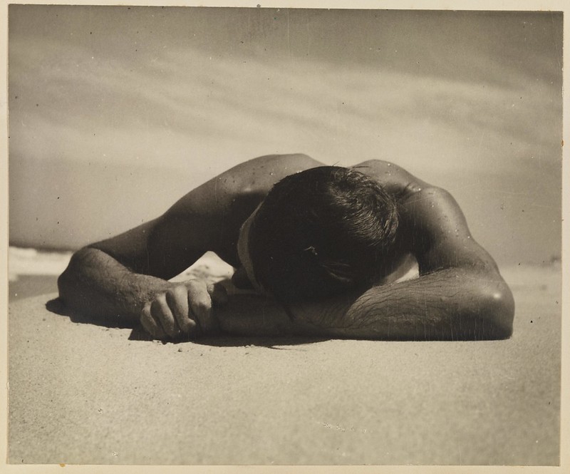 A man laying on a beac with his head in his hands, shown looking slightly downward on the top of his head