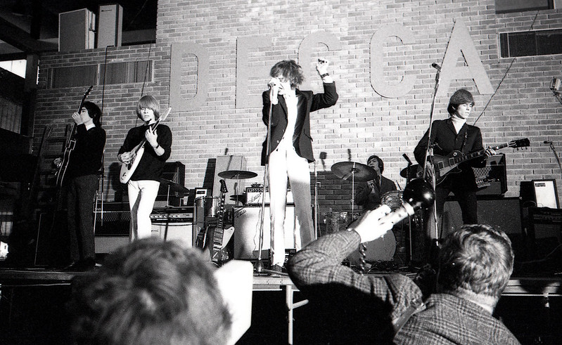 The Rolling Stones playing a concert in a small auditorium. Mick Jagger is jumping in the air and the word DECCA is on the back brick wall in sans serif letter.