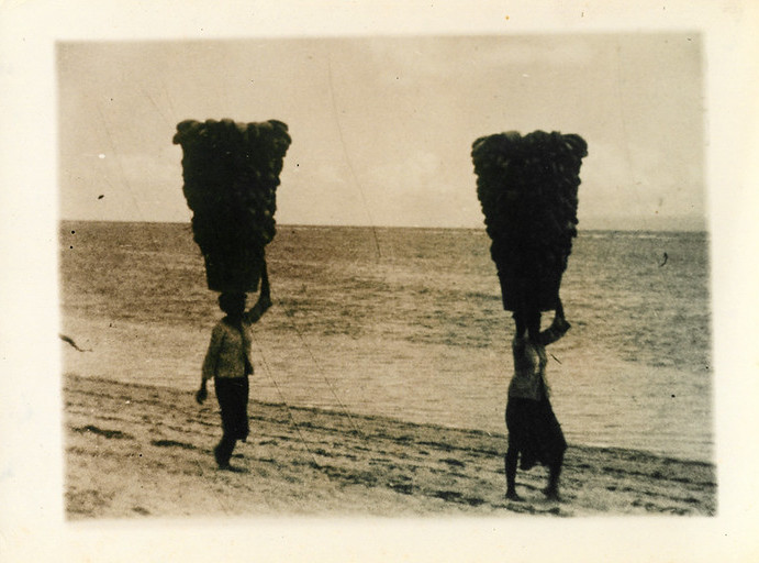 two people carrying very large baskets on their head walking along a beach