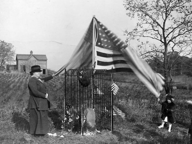 A woman stands near a rock with an american flag waving overhead.
