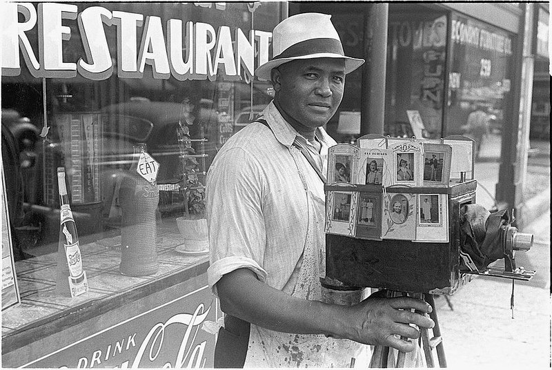 A man in a hat stands with a portable camera on which are smaller images indicating his job