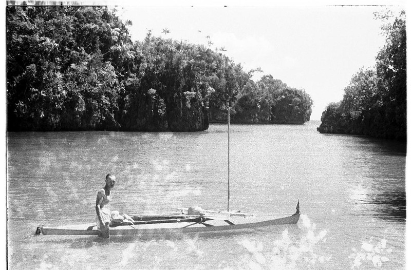 a grainy photograph of a man standing by his kayak which is in the water in a tropical location