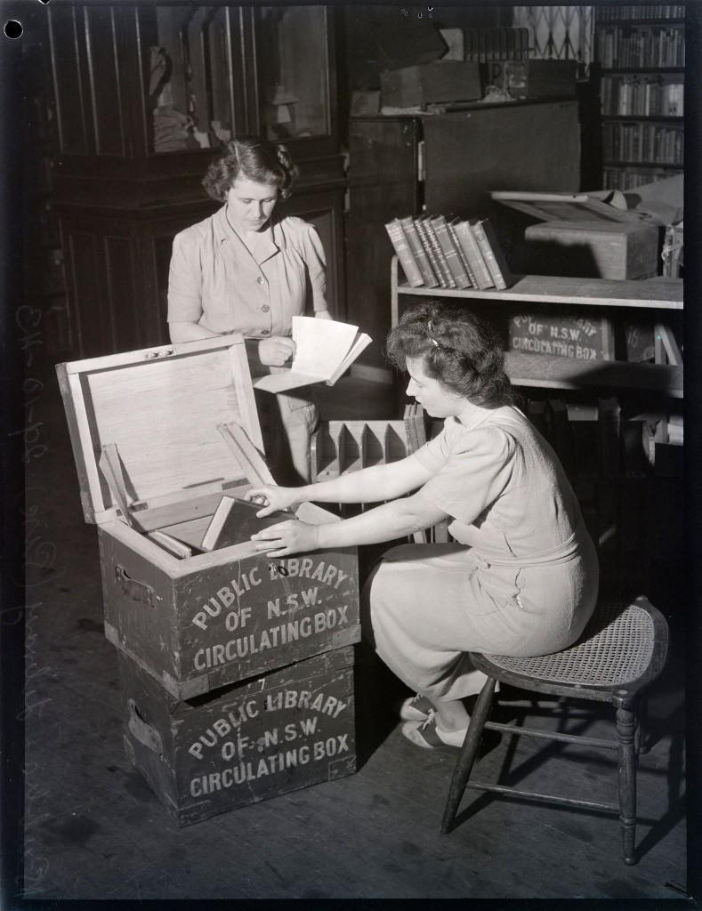 two women examine and put books into a box that says State Library of New South Wales Circulating Box