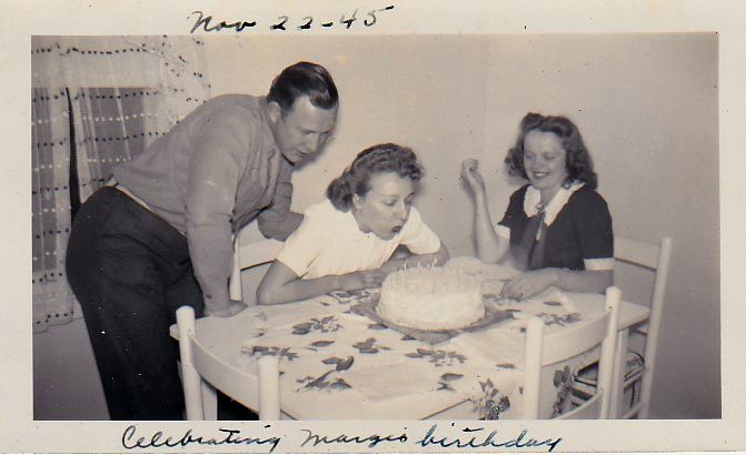 Black and white photograph featuring a white woman blowing out candles on a birthday cake. She is surrounded by another seated woman and a man, standing behind her. On the photo is handwritten 'Nov 22-45' and 'Celebrating Marge's birthday'.