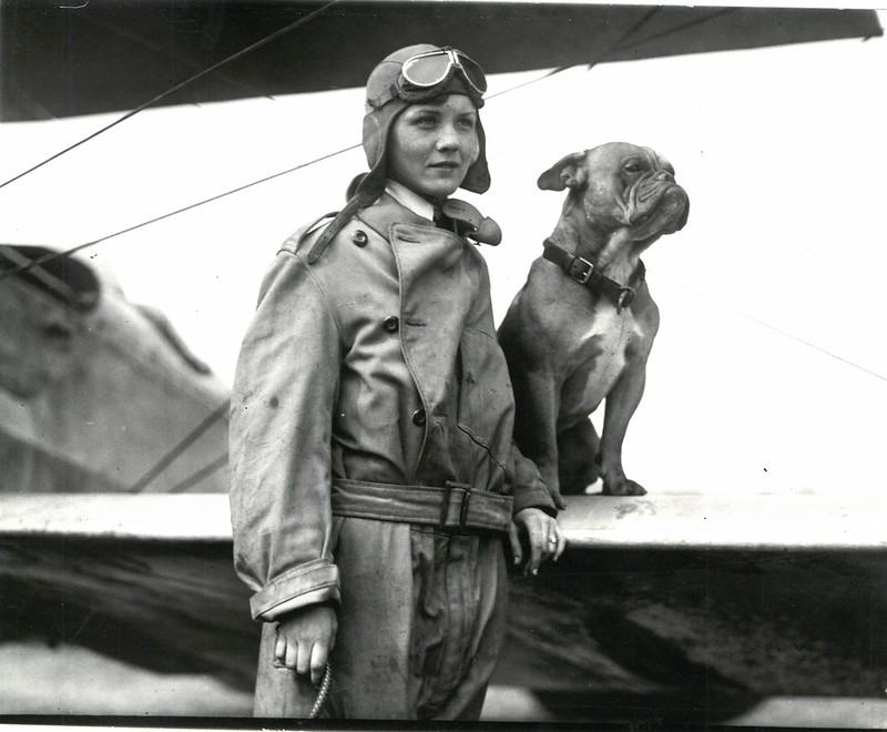 a woman in a flight outfit standsby a small airplane. There is a medium sized dog sitting on the wing of the airplane next to her