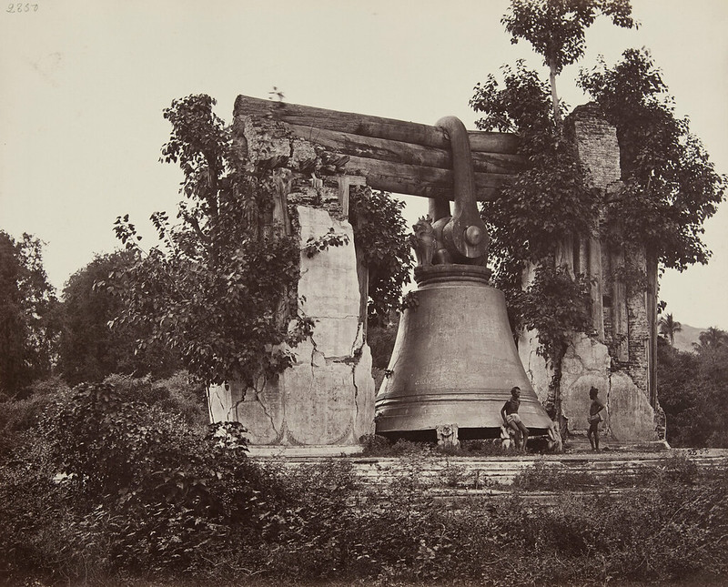 Two men standing by a bell which is significantly larger than either one of them, in a jungle setting