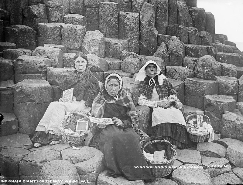 Three women sitting on Fionn Mac Cumhaill's (Finn Mac Cool) Wishing Chair at the Giant's Causeway, Co. Antrim. They're selling tourist trinkets and books containing views of the locality. The woman on the left is barefoot.