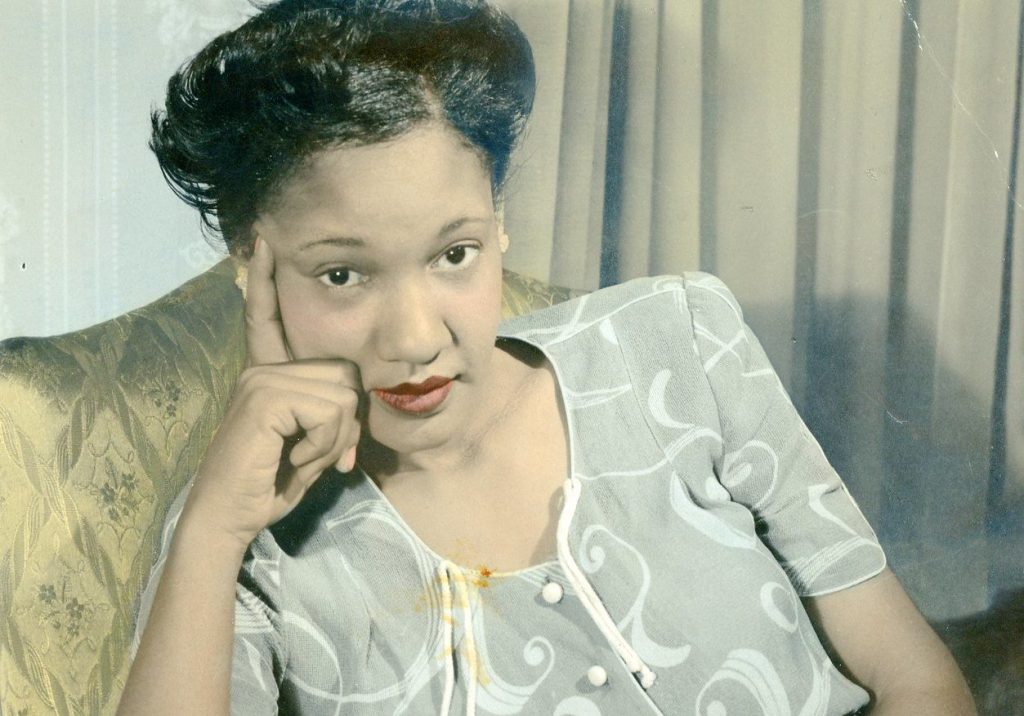 color photograph of a black woman in a print dress holding a book looking up at the photographer