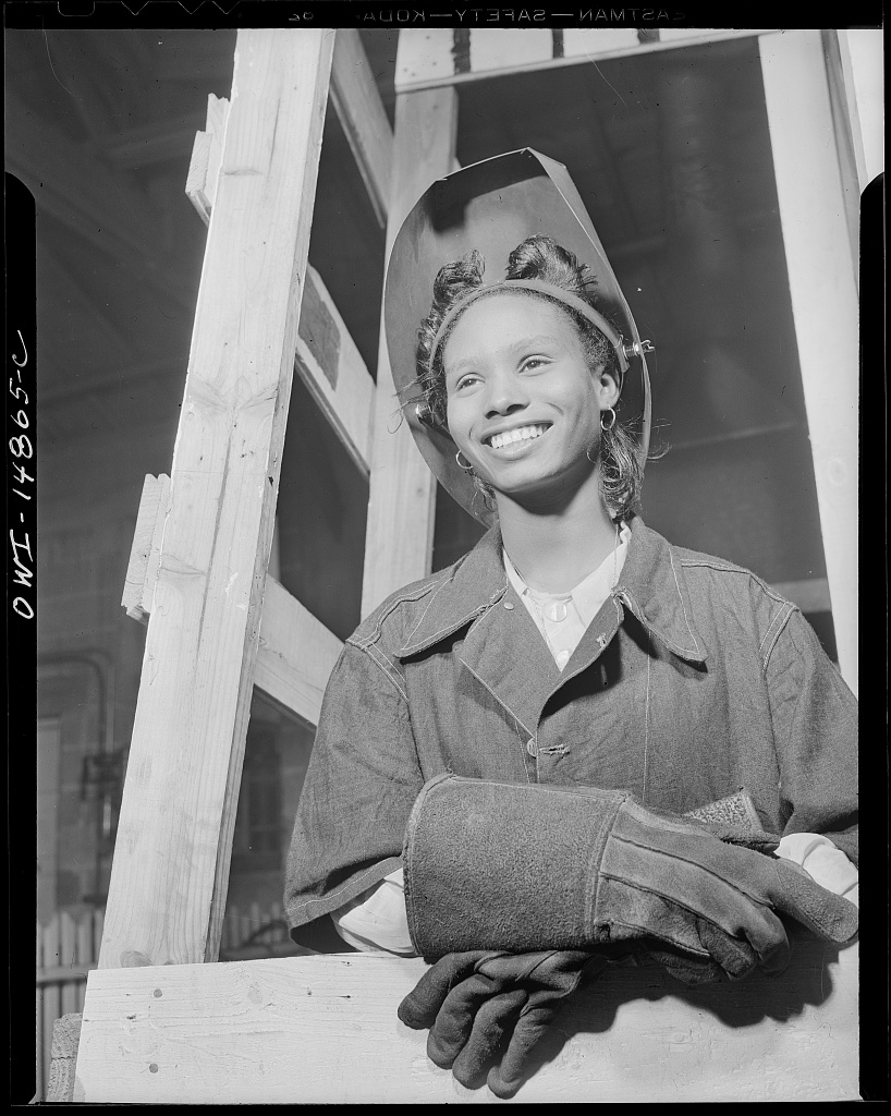 a smiling young Black woman with short hair wears a welding visor pushed back in her head and welding gloves