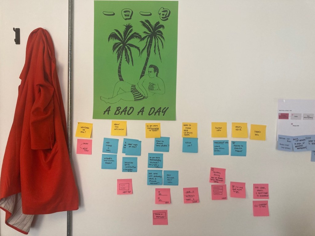 Sticky notes and a green poster on an office wall. The poster reads 'A Bao a Day' and features a drawing of a man in swim trunks drinking from a pineapple. A bao a day clearly leads to a good life.