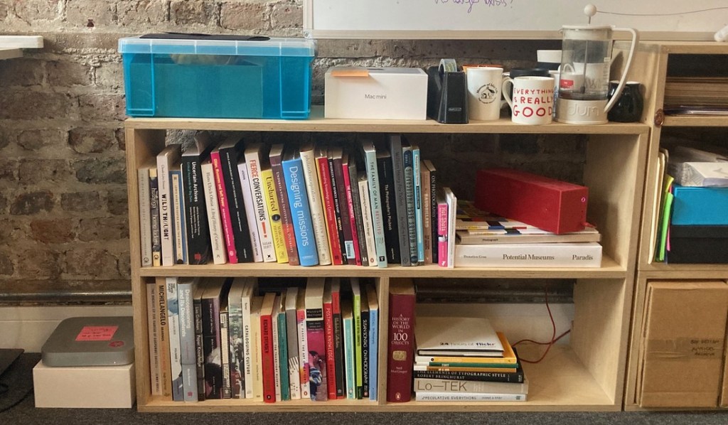 A bookshelf holds a small collection of art, tech and theory books. On top of the shelf is a coffee pot and mugs.