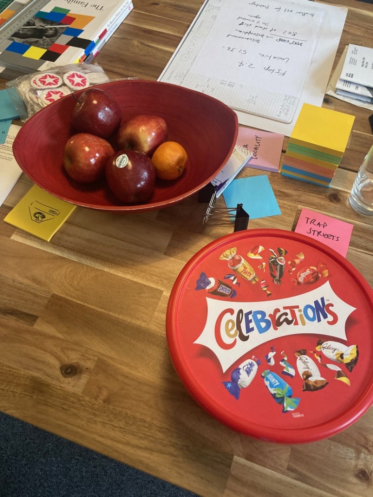 A red plastic box of Celebrations chocolates next to a fruit bowl with apples
