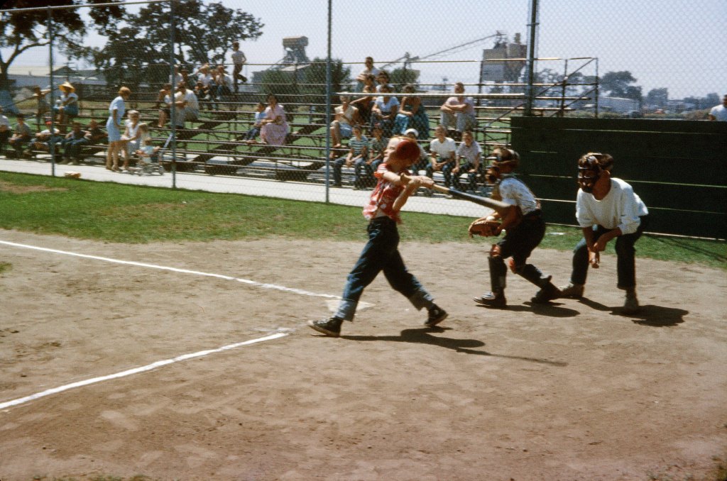 A child swinging at a baseball in a small baseball field while people watch from the stands.
