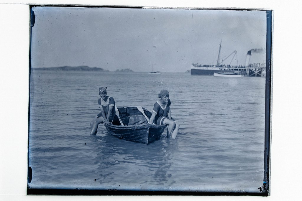 Glass plate negative showing two girls sitting on side of a dinghy on the water